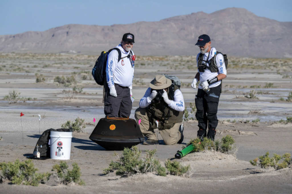 Humans dressed in white hazmat suits look at object.
